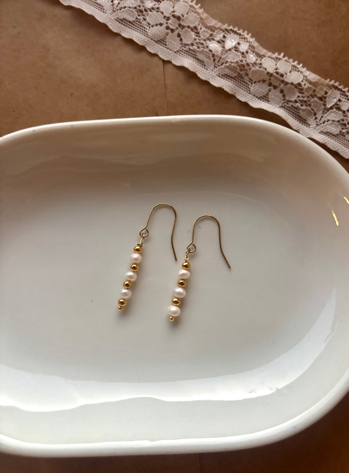 A pair of gold dangle earrings with small white pearls, displayed on a white ceramic dish over a brown surface with a lace ribbon in the background.