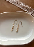 A pair of gold dangle earrings with small white pearls, displayed on a white ceramic dish over a brown surface with a lace ribbon in the background.