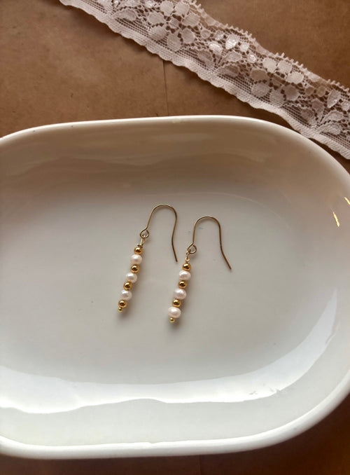 A pair of gold dangle earrings with small white pearls, displayed on a white ceramic dish over a brown surface with a lace ribbon in the background.